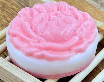 May include: A round, pink and white soap bar shaped like a peony flower, resting on a small wooden soap dish. The soap has a detailed floral design with a light pink color and a white base.