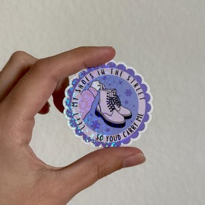 May include: A round sticker with a scalloped edge and a glittery border. The sticker features a pair of light purple boots with white laces and the text "Lift my shoes in the street so you'd carry me."