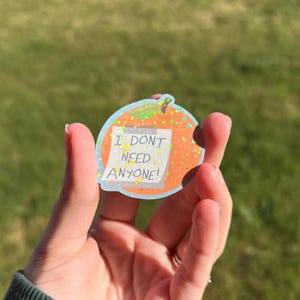 May include: A glittery orange sticker in the shape of a fruit with a light blue outline. The sticker has a white sticky note with the text "I DON'T NEED ANYONE!" in black letters.