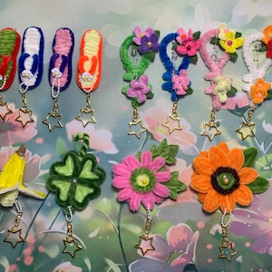 Peut inclure: Une collection de porte-clés faits à la main avec divers motifs. Les porte-clés comprennent des tongs colorées, des formes florales, une banane, un trèfle à quatre feuilles et des fleurs. Chaque porte-clés a un charme en forme d'étoile dorée.