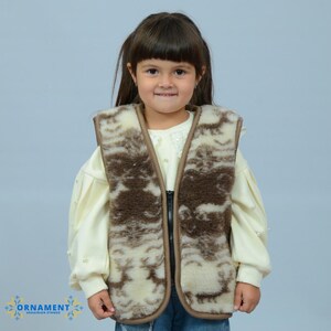 Könnte beinhalten: Eine Kinderweste aus cremefarbenem und braun gemustertem Material. Die Weste hat eine braune Einfassung und einen Reißverschluss. Das Kind trägt ein cremefarbenes Oberteil und blaue Jeans. Die Weste scheint aus einem weichen, möglicherweise wollenen Material zu sein.