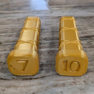 Może przedstawiać: Dwa złote, prostokątne obiekty o teksturowanych bokach i zaokrąglonych końcach. Jeden obiekt ma numer 7, a drugi numer 10. Obiekty są prawdopodobnie częścią zestawu lub narzędziem pomiarowym.