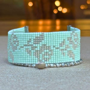 May include: A wide cuff bracelet crafted from light mint green seed beads featuring a repeating silver bead pattern of floral motifs. The lower edge is accented with a single row of metallic silver beads and a small, centered silver charm.