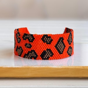 May include: A cuff bracelet crafted from small, square beads in a vibrant neon orange colour. The bracelet features a repeating pattern of dark brown and black geometric shapes that resemble an animal print design across the entire surface.