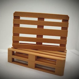 Könnte beinhalten: Zwei kleine, hellbraune Holzpaletten. Eine Palette steht aufrecht, die andere liegt flach. Die Paletten haben eine strukturierte Oberfläche und sind mit horizontalen Latten und vertikalen Stützen versehen, die Miniatur-Versandpaletten ähneln.