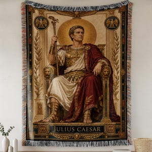 Op de afbeelding: Geweven wandtapijt met een portret van Julius Caesar op een troon. Het kunstwerk toont Caesar in een gouden borstplaat, witte toga en rode mantel, met een staf. Het wandtapijt heeft een gefranjerde rand en de tekst "JULIUS CAESAR".