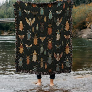 May include: A dark blanket featuring a repeating pattern of various insects in shades of brown, blue, and beige. The blanket has a fringed edge and is held up by a person standing in water.