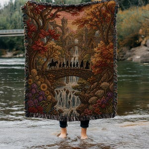May include: Woven tapestry blanket featuring a fantasy landscape with a bridge, waterfalls, and figures. The blanket has a fringed edge and depicts a scene with trees, flowers, and a castle in warm autumnal colors. The blanket is held up by a person standing in water.
