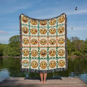 May include: A woven blanket featuring a repeating pattern of pixelated emoji faces in various expressions. The blanket has a cream-colored background with green and orange square borders. The fringe is a mix of colors. The blanket is held up by a person on a wooden dock.