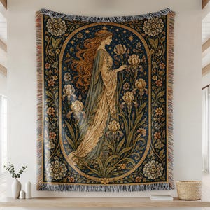 May include: A woven tapestry throw blanket featuring a central oval frame containing a figure with long flowing hair standing among iris flowers and stars against a dark blue background, surrounded by a floral border with fringed edges.