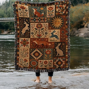 Könnte beinhalten: Eine gewebte Wohndecke mit einem mehrteiligen Design, das Illustrationen von Füchsen, Hasen, Blumen und einer Sonne zeigt. Das Textil im Wandteppich-Stil hat einen Fransenrand und eine Farbpalette aus Dunkelrot, Orange, Blau und Creme.