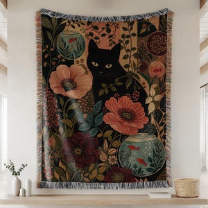 以下が含まれることがあります： 黒い猫が中央に描かれ、その周りに大きな赤やピンクの花、緑の葉、そして小さな赤い魚が入った2つの丸い金魚鉢が配置された織物のタペストリー風ブランケット。長方形の布地は、上下の端にフリンジが付いています。