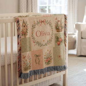 May include: A woven throw blanket featuring a patchwork design with floral motifs, a birdhouse, a basket of flowers, strawberries, and bows. The center panel displays the name Olivia inside a floral wreath with a pink bow. The edges have fringe.