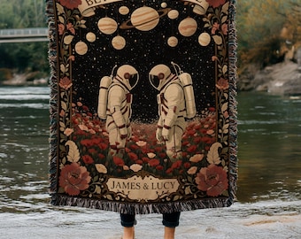 May include: A woven throw blanket featuring a space-themed design. The black background displays planets and two astronauts in white space suits standing in a field of red flowers. The words "BEYOND UNIVERSE" and "JAMES & LUCY" are also visible.