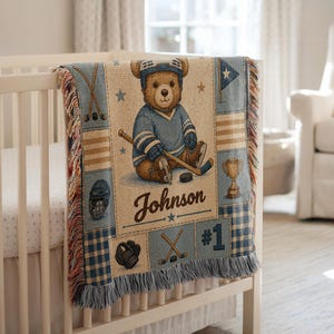 May include: A woven throw blanket featuring a brown teddy bear wearing a blue hockey uniform, helmet, and skates. The design includes hockey sticks, a puck, a trophy, and the name Johnson with the number 1. The blanket has a fringed edge.
