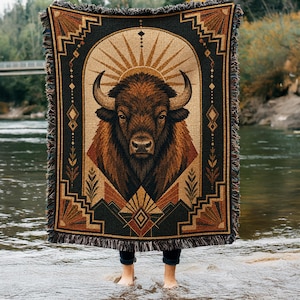 May include: A rectangular woven throw blanket featuring a central illustration of a brown bison head framed by an archway. The design includes geometric patterns, sunburst motifs, and botanical elements in shades of black, tan, and rust with fringe.