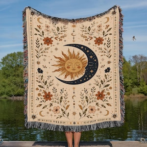 May include: A woven throw blanket featuring a central sun and crescent moon design in black and gold tones. The cream-colored fabric is decorated with botanical floral patterns, stars, and small moon phases, finished with a multi-colored fringe edge.