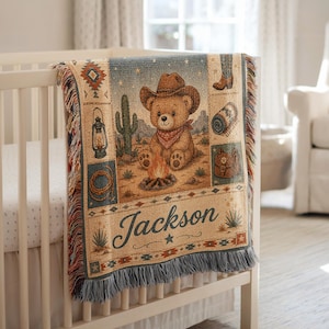 May include: A woven throw blanket featuring a central illustration of a teddy bear wearing a cowboy hat and bandana sitting by a campfire. The design includes a cactus, lantern, lasso, and boots, with the name Jackson printed in dark blue script.
