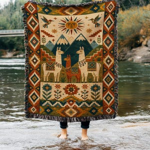 May include: A woven throw blanket featuring a central design of three llamas standing before mountains and a sun. The border displays geometric patterns in orange, teal, and cream. The blanket has a fringed edge and a colorful, detailed illustration.