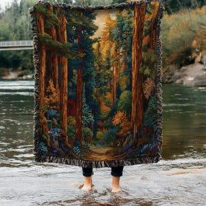 Puede incluir: Una manta tejida rectangular que presenta un paisaje forestal detallado con troncos de árboles altos de color marrón, follaje verde denso y luz dorada filtrándose a través del dosel. La manta tiene un borde con flecos decorativos.