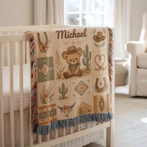 Peut inclure: Un plaid tissé portant le nom Michael, un ours en peluche avec un chapeau de cowboy, des cactus, des fers à cheval, un lasso et un crâne de vache. La couverture a un fond crème avec des motifs multicolores et des franges bleues en bas.