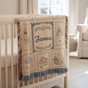 Puede incluir: Una manta tejida con un diseño de sello postal con el nombre James en el centro. La manta muestra ilustraciones de un biberón, un arcoíris, una nube durmiente, zapatos de bebé y un conejo en una cesta con flecos en los bordes.