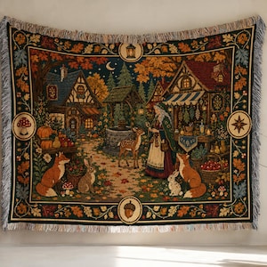 May include: A woven tapestry blanket featuring a detailed autumn forest scene with a cottage, a stone well, a person in a cloak, a deer, foxes, and rabbits. The border is decorated with oak leaves, acorns, mushrooms, and stars on a dark background.