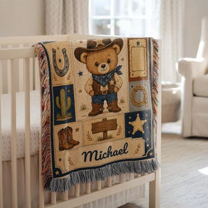 May include: A woven throw blanket featuring a central teddy bear dressed as a cowboy with a hat, vest, and boots. The design includes a horseshoe, cactus, lasso, star, and a wooden sign with the name Michael printed in dark blue script text.