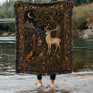 May include: A rectangular woven throw blanket featuring a dark forest scene with a large white stag, a person holding a lantern, a crescent moon, and various woodland animals including rabbits and a fox, finished with a decorative fringed border.