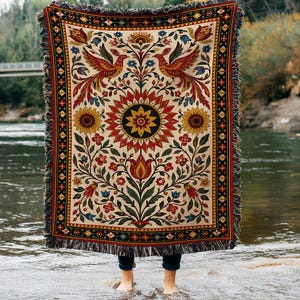 May include: A woven throw blanket featuring a cream background with a symmetrical pattern of two red birds, a large central sunflower, and various floral motifs in red, yellow, green, and blue. The blanket has a decorative border and fringed edges.