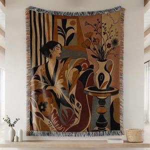 Op de afbeelding: Een geweven wandtapijt met een persoon die in een stoel zit naast een bijzettafel met een vaas met bloemen. Het ontwerp gebruikt een warm kleurenpalet van bruin, beige en zwart met abstracte vormen, verticale strepen en franjes.