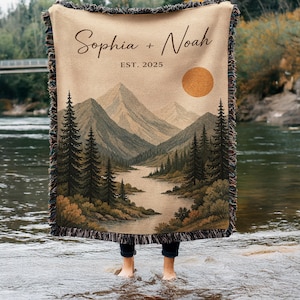 Puede incluir: Una manta tejida con bordes de flecos que presenta una ilustración de un paisaje con montañas, un río y pinos. El texto "Sophia + Noah" y "EST. 2025" está impreso en la parte superior, junto a un gran sol naranja sobre un fondo beige.