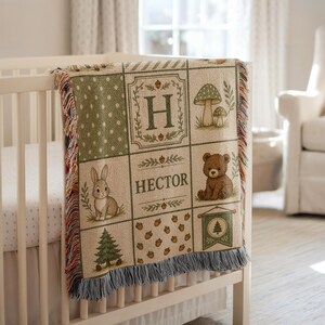 May include: A woven throw blanket featuring a grid pattern with illustrations of a rabbit, a bear, mushrooms, pine trees, and acorns. The center panel displays the letter H and the name HECTOR in green text. The blanket has a fringed edge.