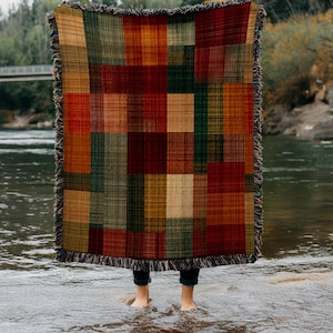 May include: A woven throw blanket with a patchwork design in shades of red, green, gold, and brown. The blanket has a fringed edge and is held up in front of a person standing in water.