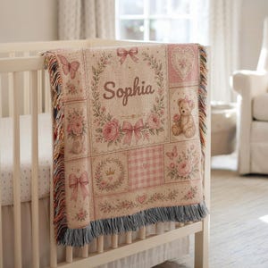 May include: A woven throw blanket featuring a patchwork design in shades of pink and cream. The blanket displays the name Sophia, a teddy bear, floral wreaths, butterflies, hearts, bows, and a crown, finished with a fringed edge on the bottom.