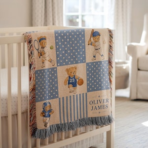 May include: A woven throw blanket featuring a grid pattern of teddy bears playing various sports including tennis, baseball, and basketball. The blanket has blue and white polka dot and striped sections, with the name OLIVER JAMES printed on it.