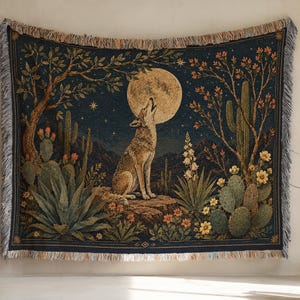 May include: A woven tapestry blanket featuring a dark blue night sky with a large full moon and a coyote howling. The scene includes desert flora such as cacti, agave, and flowering shrubs. The rectangular textile has a fringed edge border.