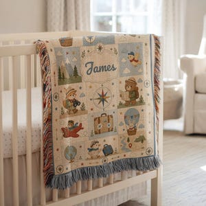 May include: A woven throw blanket featuring a grid pattern with illustrations of bears, a rabbit, a bird, mountains, a compass, a suitcase, and a globe. The name James is printed in the centre. The blanket has a fringed edge in blue and tan.