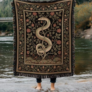 May include: A black woven blanket with a detailed design of a snake entwined around a tree, surrounded by roses and foliage. The blanket has a fringed edge and is held up in front of a river.