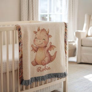 Peut inclure: Une couverture de bébé beige avec une illustration de dragon de dessin animé. Le dragon est orange clair avec un visage souriant et les bras tendus. La couverture a une bordure à franges multicolores et le nom "Robin" est imprimé.