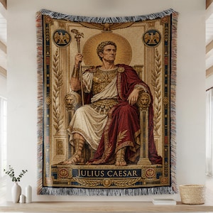 May include: A woven tapestry throw blanket featuring a depiction of Julius Caesar seated on a throne, wearing armor and a red cloak, holding a staff. The design includes gold, red, and blue colors with laurel motifs and the text JULIUS CAESAR.