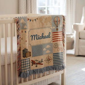 May include: A woven throw blanket featuring a travel-themed patchwork design with the name Michael in blue script. The pattern includes a hot air balloon, a vintage airplane, suitcases, a compass, a world map, and postage stamps in muted tones.
