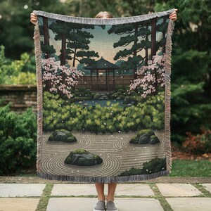 Puede incluir: Una manta tejida con una escena de jardín japonés con una pagoda, un estanque y flores de cerezo. La manta tiene un borde con flecos y representa un paisaje sereno con árboles, rocas y un jardín de arena rastrillada. La paleta de colores incluye verdes, azules y rosas.
