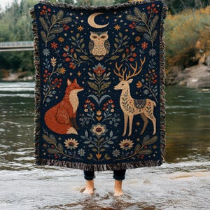 May include: A dark blue woven throw blanket featuring a symmetrical pattern of a central owl, a fox on the left, and a deer on the right. The design includes floral motifs, pine branches, stars, and a crescent moon, finished with a fringed edge.