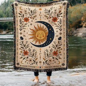 May include: A woven throw blanket featuring a central sun and crescent moon design in gold and dark blue. The cream-colored fabric is decorated with botanical floral patterns in orange, green, and blue, finished with a fringed edge on all sides.
