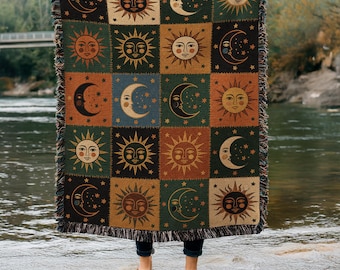 May include: A woven throw blanket featuring a grid pattern of celestial motifs including suns and crescent moons with faces. The blanket has a fringed edge and is decorated in a color palette of dark green, burnt orange, black, and cream tones.