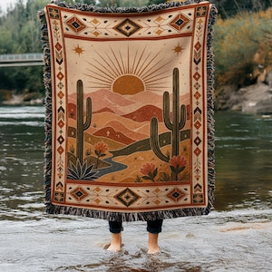 May include: A woven throw blanket featuring a desert landscape with a rising sun, saguaro cacti, and blooming flowers in shades of orange, brown, and cream. The blanket has a decorative geometric border and a fringed edge on all four sides.