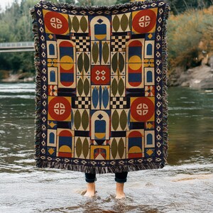 May include: A woven throw blanket featuring a symmetrical geometric pattern in red, blue, yellow, olive green, and cream. The design includes circles, arches, and checkered squares with a fringed edge along the top and bottom borders.