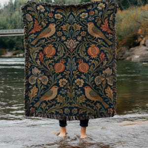 May include: A dark blue woven throw blanket with a repeating pattern of birds, pomegranates, and floral elements in shades of brown, orange, and green. The blanket has a fringed edge and is held up by a person standing in water.