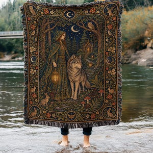 May include: A woven throw blanket featuring a detailed tapestry design of a hooded figure holding a lantern and standing beside a wolf in a forest. The scene includes a crescent moon, stars, owls, rabbits, and floral borders with fringed edges.
